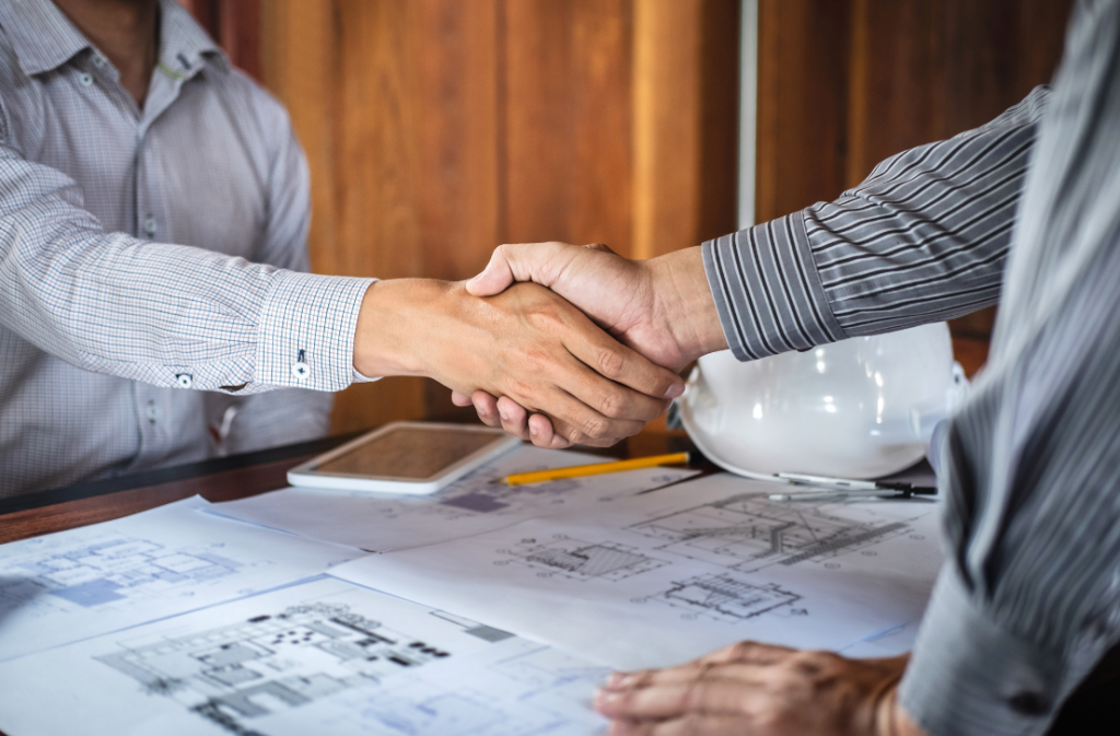 Two men shaking hands over blueprints with a hard hat, tablet and drawings on the table.