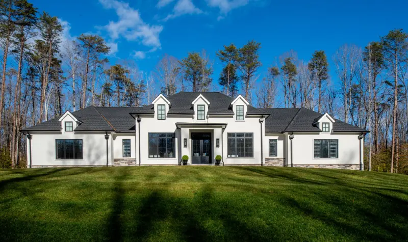 Wide front view of a luxury French Country style home with white stucco walls, dormers, black-framed windows, stone accents, and a spacious single-level living design.